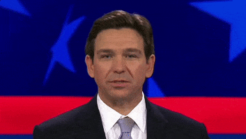 Video gif. Ron DeSantis gives a slight smile as he flicks his tongue out to touch his top and bottom teeth in front of a patriotic background. 