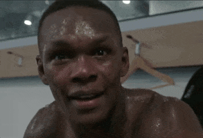Sports gif. Izzy, aka Israel Adesanya, a UFC fighter, is shirtless and sweating profusely after a workout. He looks at us with a curious smile and gently pushes away the camera.