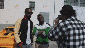 Celebrity gif. Kanye West wears a pair of sunglasses while being recorded outside. He sees us out of the corner of his eye and cocks his head to flash us an unexpectedly wide grin.