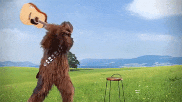 Star Wars gif. Chewbacca appearing in a spacious landscape, smashing an acoustic guitar on a stool.