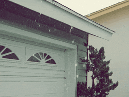 Video gif. Hail falls in front of a white garage that has a lonely tree resting next to it.