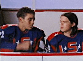 Movie gif. Elden Henson as Fulton Reed and Aaron Lohr as Dean Portman fist bump as they sit on the sidelines of the ice. 