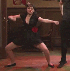 TV gif. A young woman, wearing a black dress with a red flower ornament on her hip and a feathery hair decoration, does a provocative modern dance. Next to her, a man dressed in black does an Irish step dance.