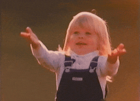 Video gif. Little girl in overalls smiles and reaches out to catch a ball. The ball hits her chest as she moves in to grab it, but her arms are too far up and the ball falls on the ground.