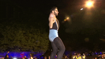 Video gif. Man wearing black bodysuit and tights, denim shorts, and rainbow suspenders dances flamboyantly while police lights flash below.