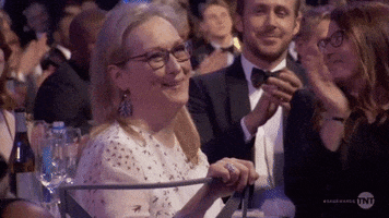 Celebrity gif. Meryl Streep smiles and blows a kiss as celebrities in formal wear applaud around her.