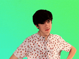 Video gif. Declan McKenna stands in front of a green screen raising his hand in the air while bowing his head, giving a shy wave goodbye. 