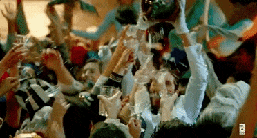 Video gif. Partygoers bounce around with martini glasses lifted in the air as a woman haphazardly pours champagne from a jug. 