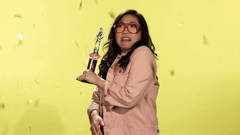 Gif of a woman holding a trophy being showered with gold confetti, at first looking shocked then waving and smiling