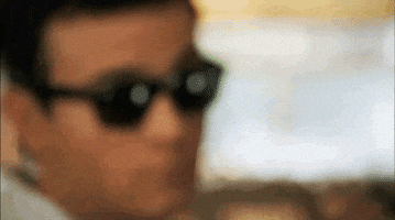 TV gif. Troy Gentile as Barry in The Goldbergs turns toward us as he comes into focus and slides his sunglasses down with a suave half smile.