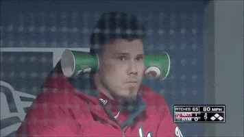 Sports gif. Jose Lobaton with the Washington Nationals Baseball team is chewing sunflower seeds and has two cups stuck on his ears to amplify the noise. He spots the camera and he holds two more cups to his eyes, looking at us.