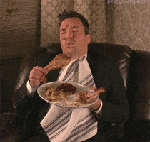 TV gif. Jimmy Fallon as host of the Tonight Show sits in a large leather recliner with a messy plate of food in his hand, some of the food smeared on his cheek and forehead. He looks at the turkey leg he's holding before groaning--his eyes are bigger than his stomach!