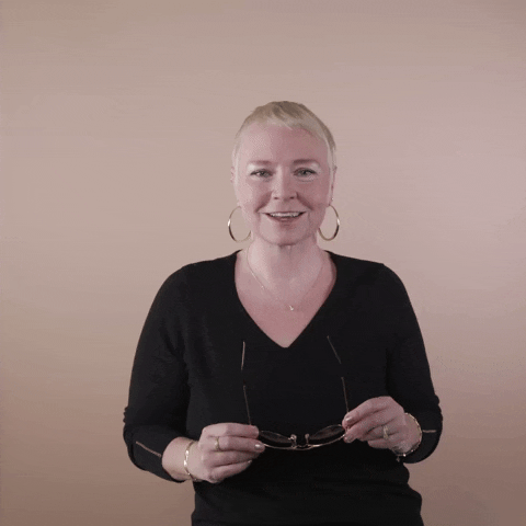 Reaction gif. A white woman with invisible disabilities, with hair styled in a sleek platinum pixie cut and big hoop earrings slides on her aviator sunglasses with a grin.
