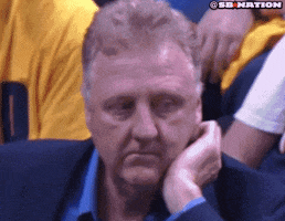 Sports gif. Larry Bird is at a basketball game looking extremely bored. He scratches his face, licks his lips, and checks his nails while spacing out.