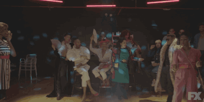 TV gif. Dyllon Burnside as Ricky on Pose wears a fur coat over his shoulders and big sunglasses. He coolly adjusts the fur coat over his shoulders as people clap for him and wave fans at him.
