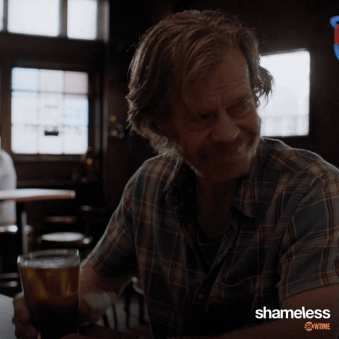 TV gif. William H. Macy as Frank in Shameless looks over toward another bar patron and raises his beer to cheers.