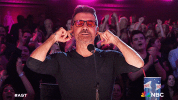 Reality TV gif. Simon Cowell in red tinted glasses on "America's Got Talent" stands at a microphone putting his fingers in his ears like he's trying to block out the noise, perhaps the happy clapping crowd behind him. 