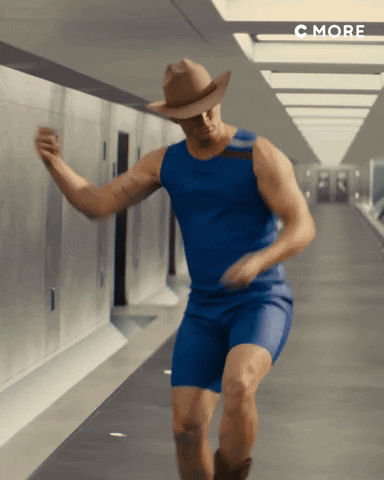 Celebrity gif. Wearing a cowboy hat, brown boots, and a blue aerobics outfit, Channing Tatum dances in a long, empty hallway.