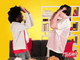 Ad gif. In a vibrant room with yellow walls, splashes of red accents, and a black sofa, two young men share a fun handshake that ends with them feeding each other a Totino's pizza roll. Two bowls with a bunch of pizza rolls sits on the table in front of them.