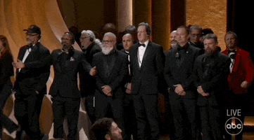 Oscars 2024 GIF. A large group of Teamsters, Gaffers, and Grips come onstage in suits and grin at the crowd as the audience cheers for them. One man wearing a headset holds his arms open wide, embracing the applause. 