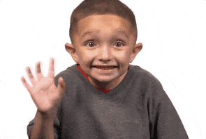Video gif. A young boy waves at us as he glances sideways and smiles sheepishly. 