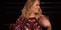 Celebrity gif. Adele turns to the side and erupts in a surprised smile as she waves off camera.