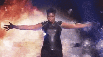 SNL gif. Leslie Jones, dressed in a shiny black top, stands in front of what looks like an explosion with her arms spread wide, threateningly staring ahead, and then dropping her arms to her side.