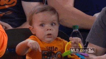 Video gif. A baby at a Baltimore Orioles game is watching the game intently and they start to shake their hands, which holds a toy, and widen their eyes as something happens on the field that excites them.