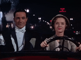 Movie gif. Gene Kelly as Don and Debbie Reynolds as Kathy in Singin in the Rain sit in a car together. With focus and a slight grin she steers the car as he glances over to her with a grin. 