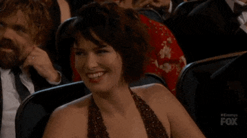 Video gif. Lena Headey at the 2015 Emmys glances over and smirks as if saying oops. 