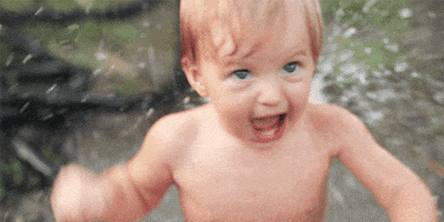 Video gif. A baby stands in front of a sprinkler flailing its short arms around. He shakes his head vigorously and screams in excitement as the water sprays all over him.