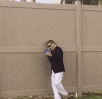 Video gif. A shaky camera takes footage of a man in a blazer and sunglasses as he speaks into a phone. He appears to be drunk: he's carrying a plastic blue cup in one hand as he stumbles backwards, and when he falls over, he spills its contents.