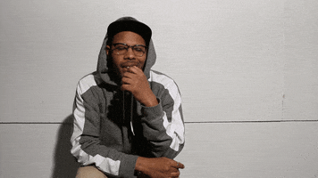 Video gif. Young man wearing a baseball cap, hoodie, and glasses, leans against a wall pensively with his hand over his mouth, nodding absently then looking at us and flashing an "ok" sign with his hand.