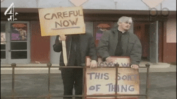 Video gif. Two priests stand in the street, holding protest signs. One says, “Careful now.” The other says, “Down with this sort of thing.”