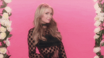 Celebrity gif. Paris Hilton flirtatiously raises a hand and waves at us in front of a pink background flanked by white roses.
