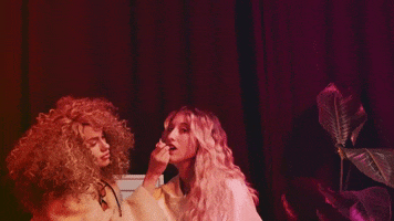 Music video gif. From Louis the Child's video for "Evalyn," a blonde wavy-haired woman sings while a brunette curly-haired woman touches up her make up, sitting in front of a red curtain next to a plant.