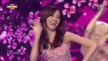 Celebrity gif. Yoona of the K-pop group Girls’ Generation waves at us during a performance of "Goodbye Stage", zooming in on her as she smiles, a large LED screen of magenta flowers in the background.