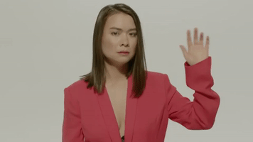 Music video gif. From the video for Your Best American Girl, Mitski, wearing an slouchy pink blazer, waves her hand, then stops and looks curiously at her hand.