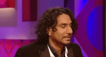 Celebrity gif. Naveen Andrews sits down and leans back, flipping his curly hair out of his face.