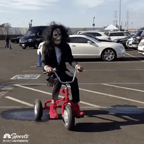 Celebrity gif. Dressed as Jigsaw from the Saw movies, Steph Curry rides a red tricycle slowly through a parking lot, glancing over his shoulder.