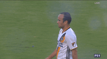 Sports gif. Landon Donovan shakes hands and hugs other soccer players on the field.