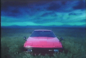 Video gif. Flashy 1980s red sports car sits alone in a vast grassy plain underneath a mysterious sky with a blanket of dark rolling clouds and a bright turquoise glow.