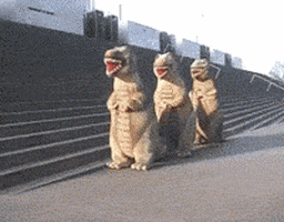 Video gif. Three people in full T-Rex costumes doing a coordinated line dance outside of an office building.