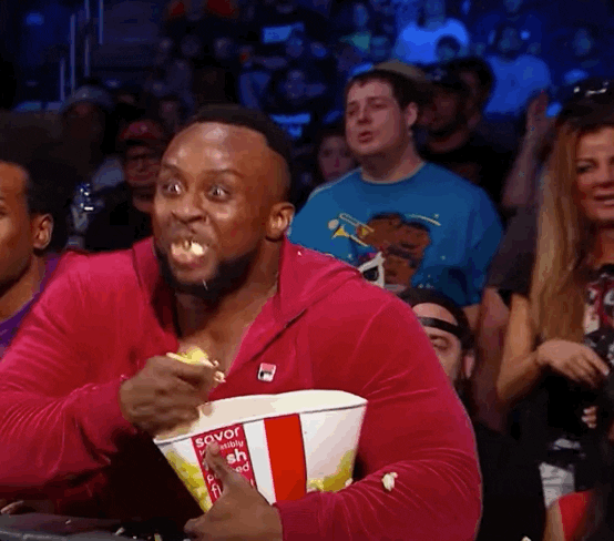 Sports gif. A WWE fan in a red hoodie stares at the ring in wide-eyed amazement as he excitedly clutches a paper bucket of popcorn.