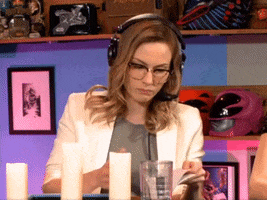 Video gif. Woman with a headset on holds up a small notebook where she has written in big letters, “Help.” She looks at us with a blank expression.