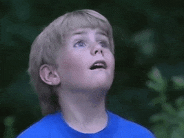 Video gif. Boy looks up and whipping his head around with awe as if something cool is moving a round in the air.