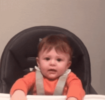 Video gif. A baby sits in a high chair and abruptly raises his fists straight up in the air with a scrunched up face.