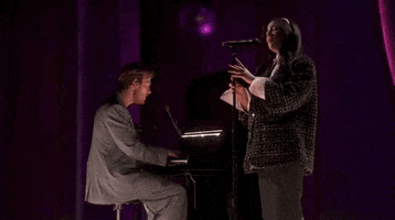 Oscars 2024 GIF. Billie Eilish and Finneas performing "What Was I Made For" on stage at the Oscars. Finneas is on the piano and Eilish stands next to him. Both are singing and Eilish waves a hand as they hit the climax of the song. 