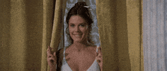 Movie gif. Mary-Margaret Humes as Miriam in History of the World: Part 1 is hiding between two curtains looks excited as she tiptoes with glee and holds both hands with crossed fingers up.