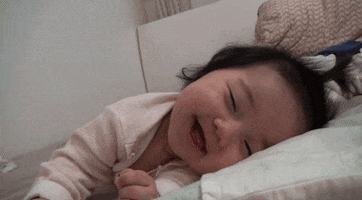 Video gif. Baby lays her head on a pillow and is at first smiling very cutely, but her tiredness catches up to her and her smile drops as she has no more energy to smile anymore.